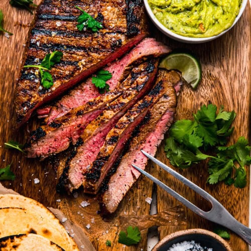 Sliced grilled carne asada on cutting board next to bowl of guacamole and cilantro leaves.