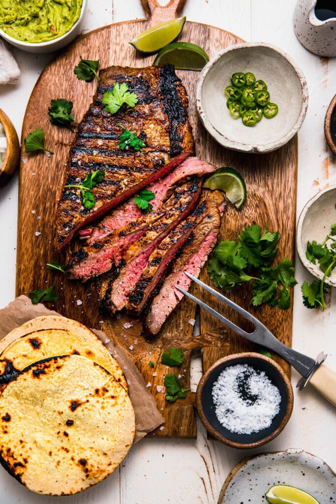 Sliced grilled carne asada on cutting board next to bowl of guacamole and cilantro leaves.