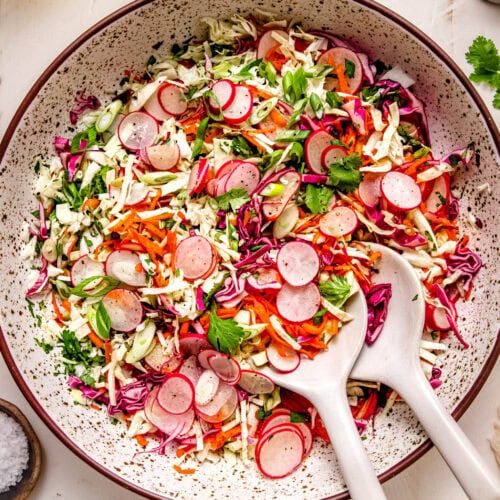 Finished radish slaw in serving bowl with spoons next to uncut radishes.