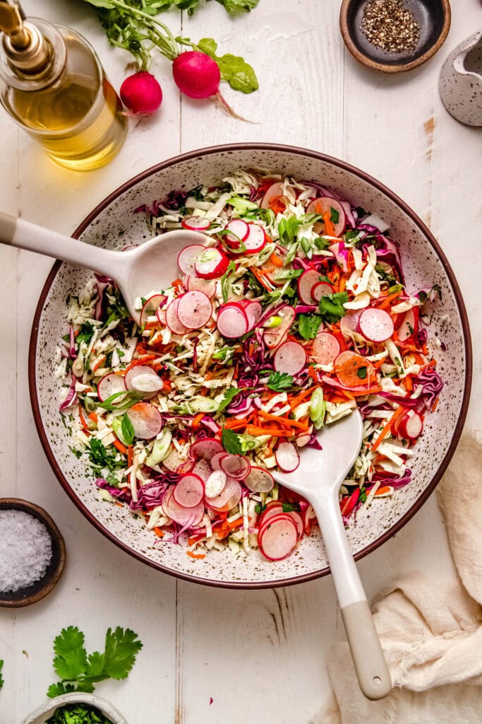 Finished radish slaw in serving bowl with spoons next to uncut radishes.