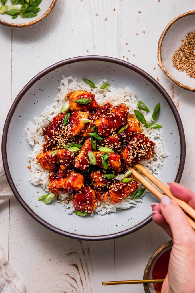 Sweet chili chicken served on bowl of rice with hand holding chopsticks.