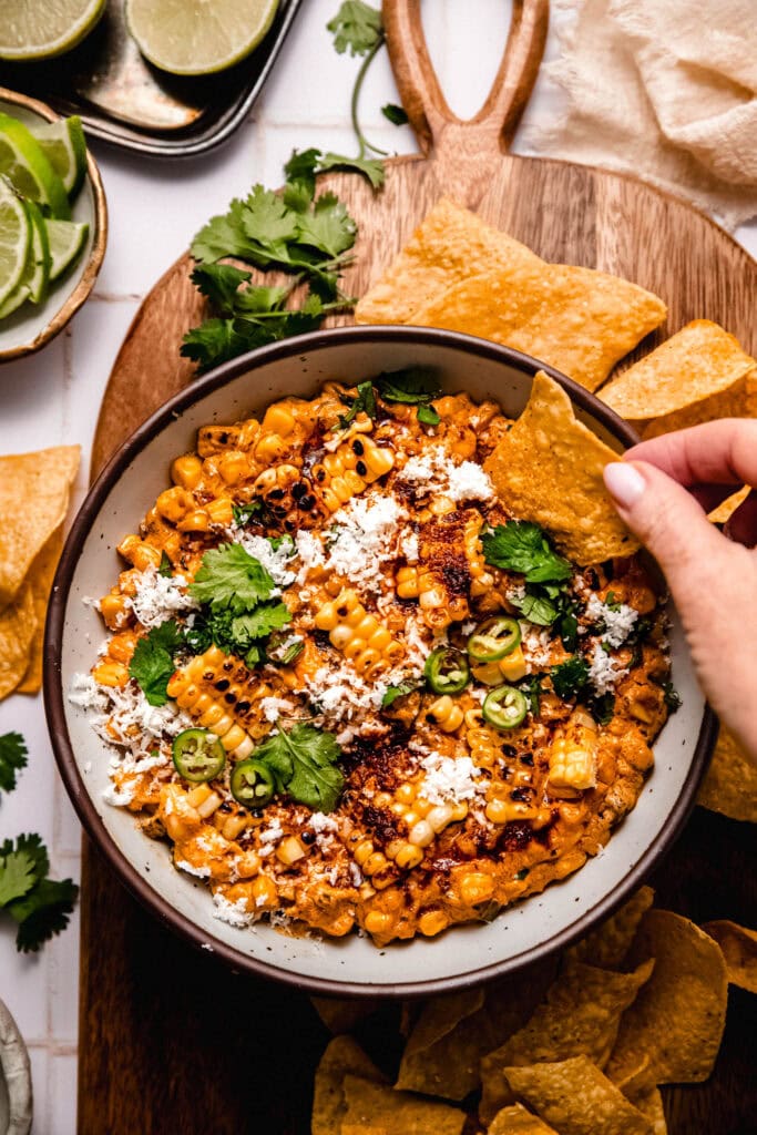 Hand dipping tortilla chip into bowl of street corn dip.