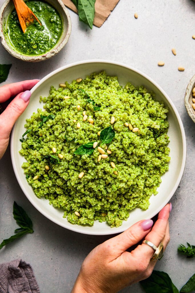 Overhead shot of hands holding bowl of pesto quinoa topped with pine nuts and basil.