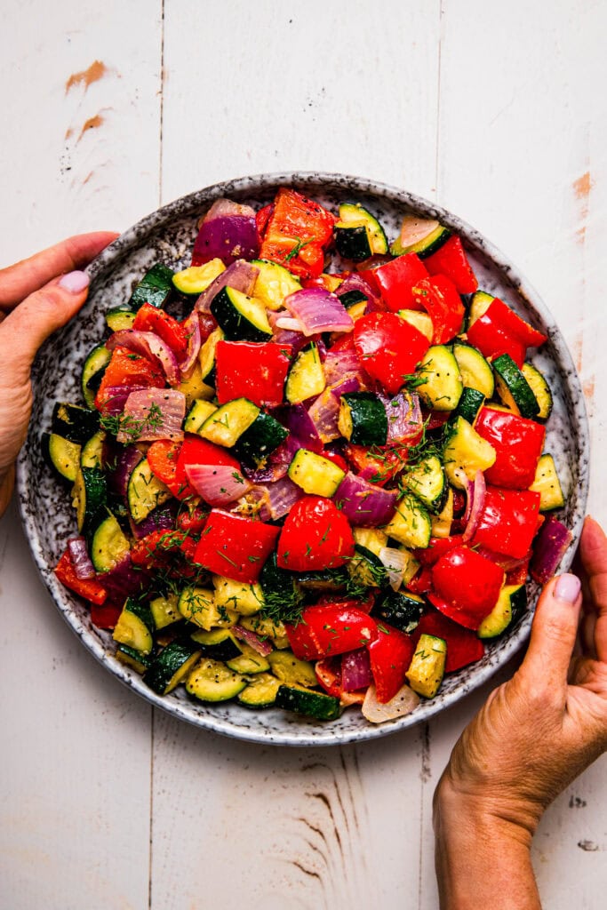 Hands holding serving plate of roasted zucchini and red peppers.