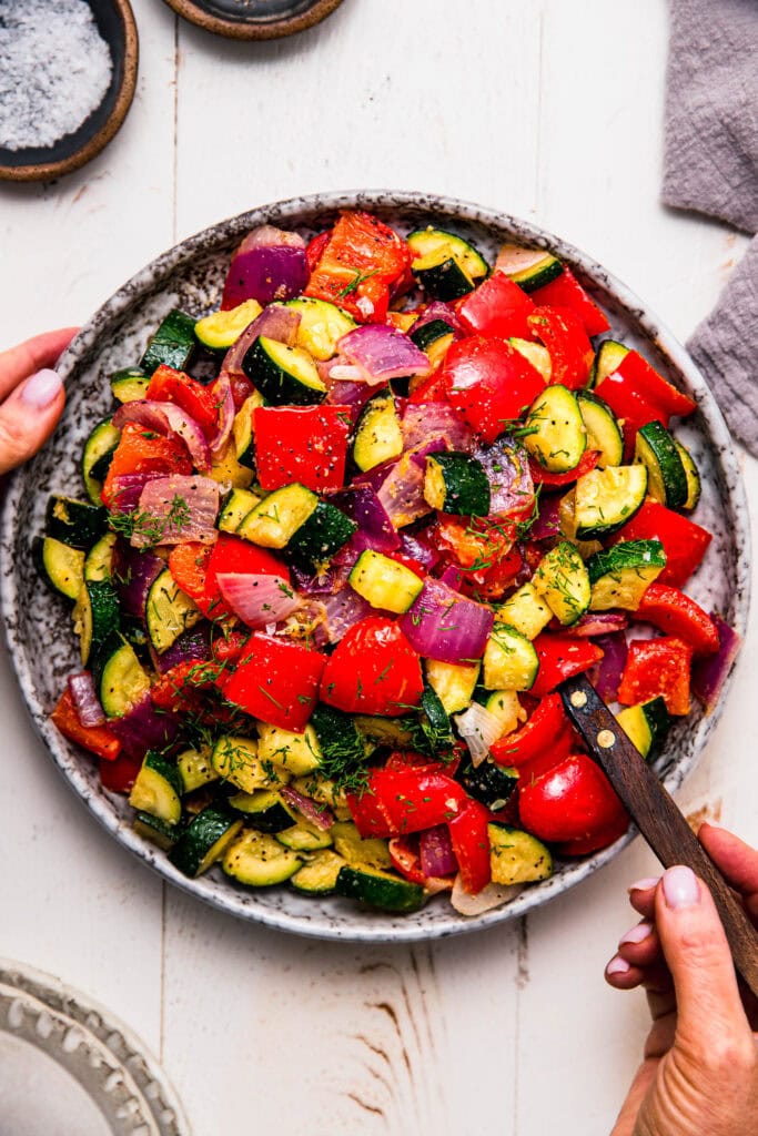 Hands holding serving plate of roasted zucchini and red peppers.