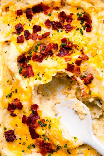 Spoon dipping into casserole dish of twice baked mashed potatoes topped with bacon & cheese.