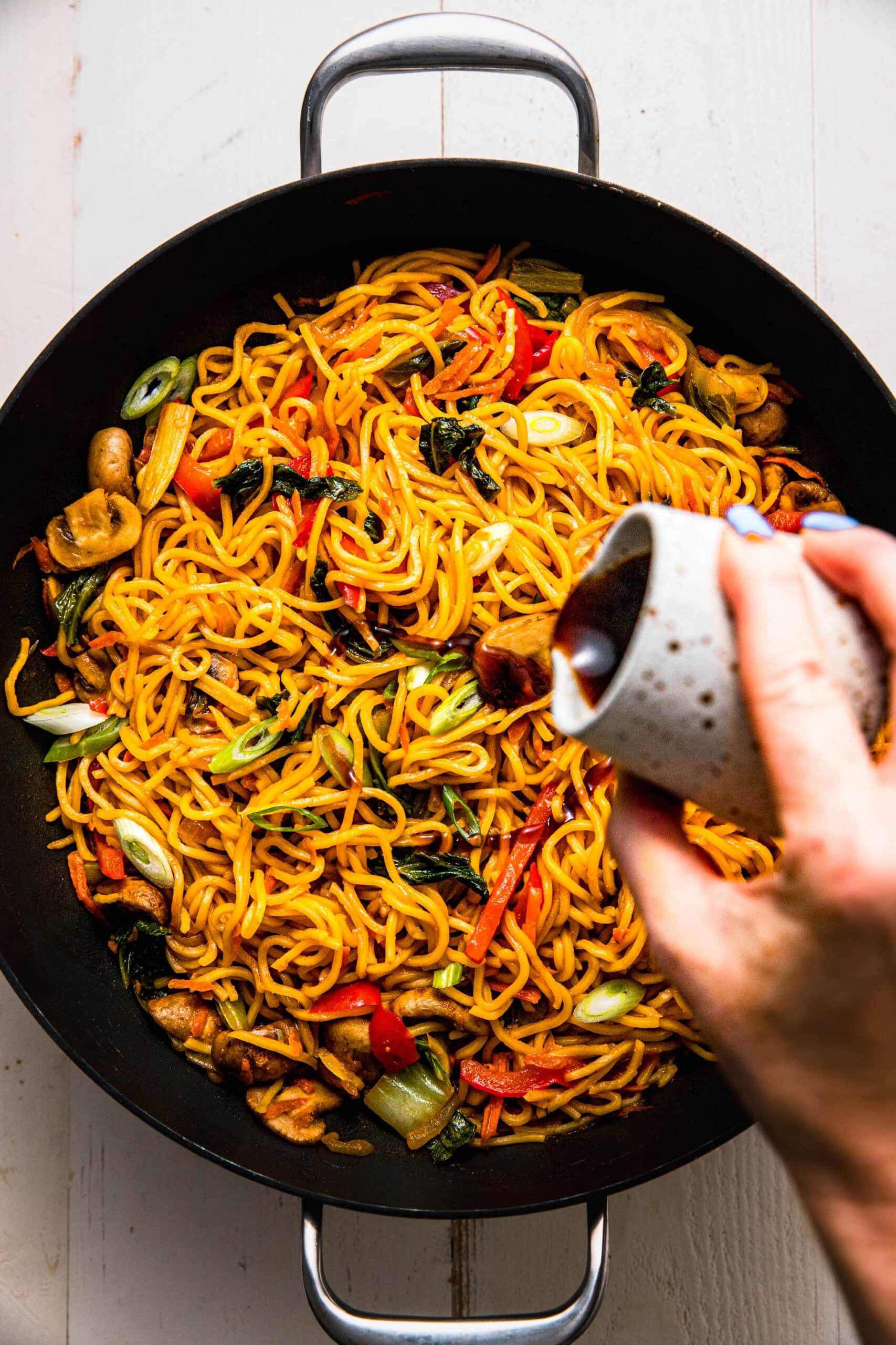 Hand pouring yakisoba sauce onto wok of cooked yakisoba noodles.
