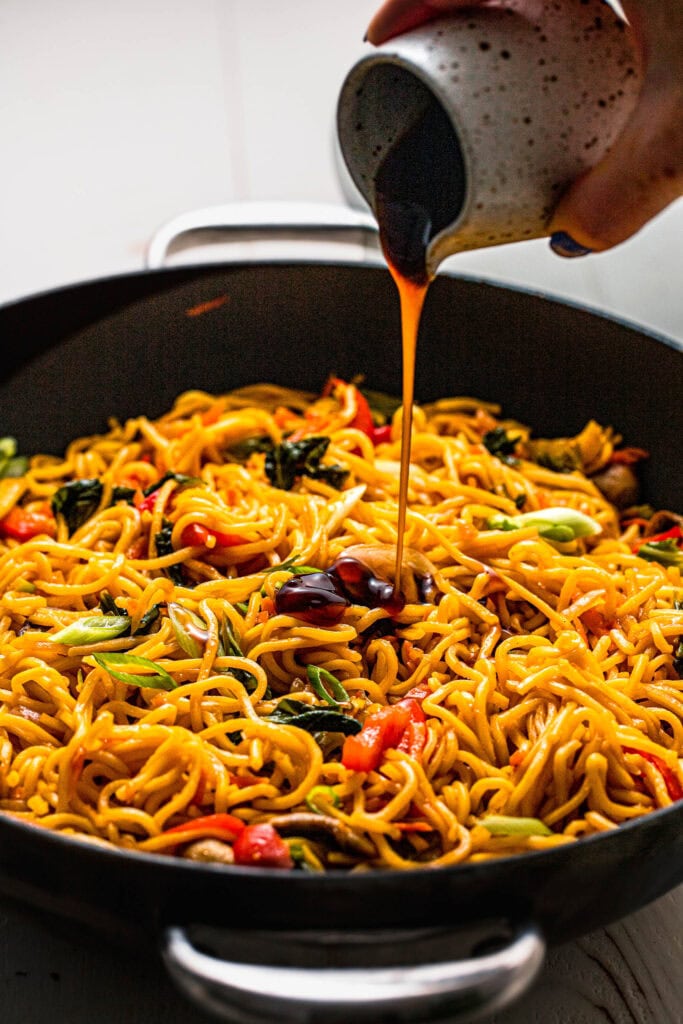 Side view of sauce being drizzled onto wok of cooked yakisoba noodles.