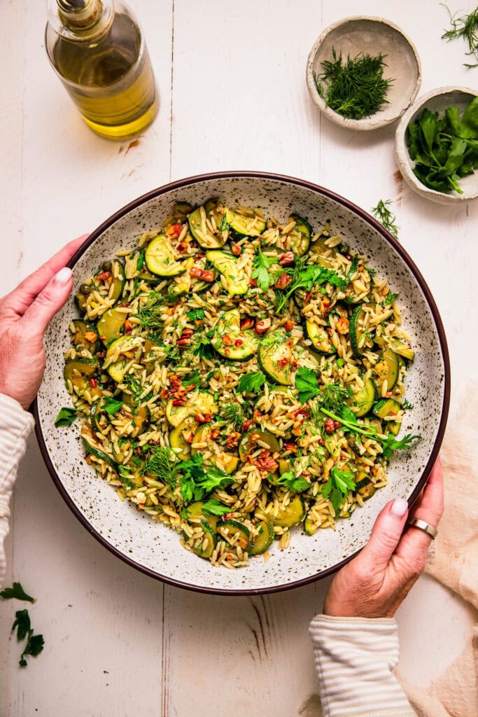 Hands holding serving bowl of zucchini orzo salad.